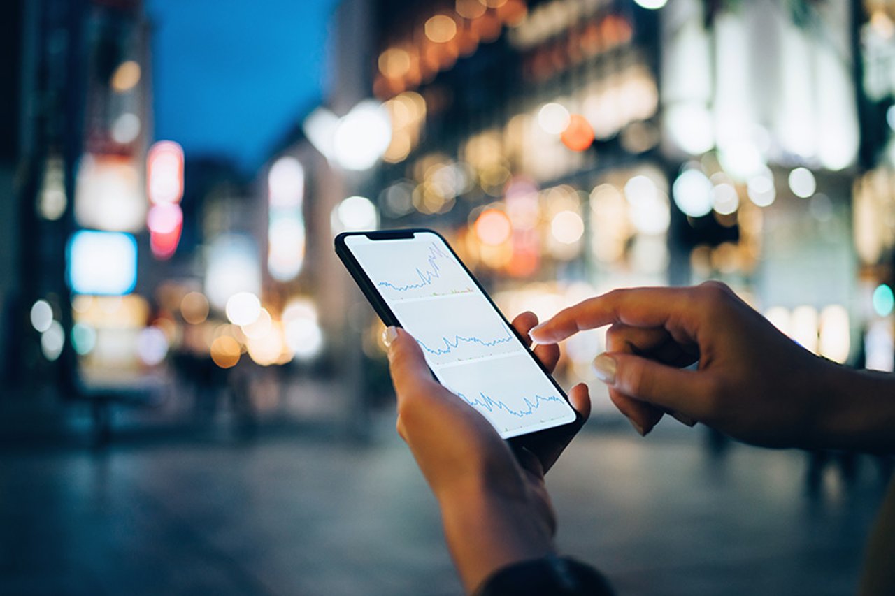 Innovative financial services companies are using communications APIs to transform the customer experience, delighting sometimes tentative customers with real-time authentication, video, voice and messaging interactions.  Businesswoman reading financial trading data on smartphone in downtown city street against illuminated urban skyscrapers